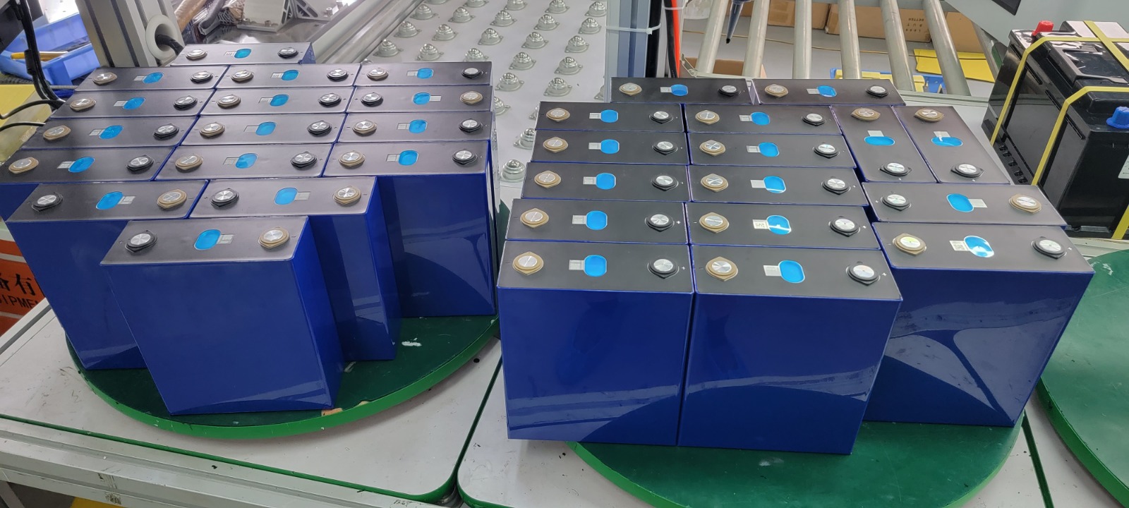 Two stacks of prismatic LFP cells on a factory work table, blue plastic cases with stainless steel tops and circular terminals — the kind that go into a self-built 16s21p pack.